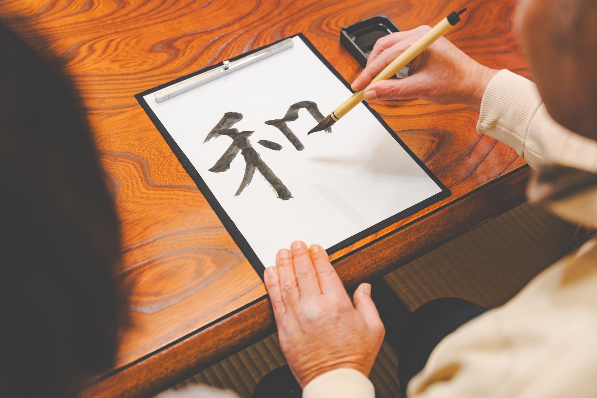 Japanese Cultural Experiences at Kyoto Abeya. Close-up of hands practicing Japanese calligraphy, focused on a white paper with the character '和' written with a calligraphy brush.