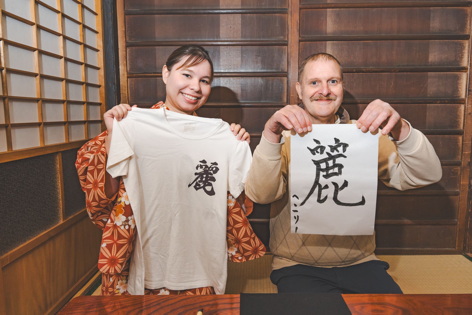 Japanese Cultural Experiences at Kyoto Abeya. A smiling man and woman each holding a T-shirt and calligraphy work with the character '愛'. Traditional Japanese room with wooden walls and shoji screens in the background.