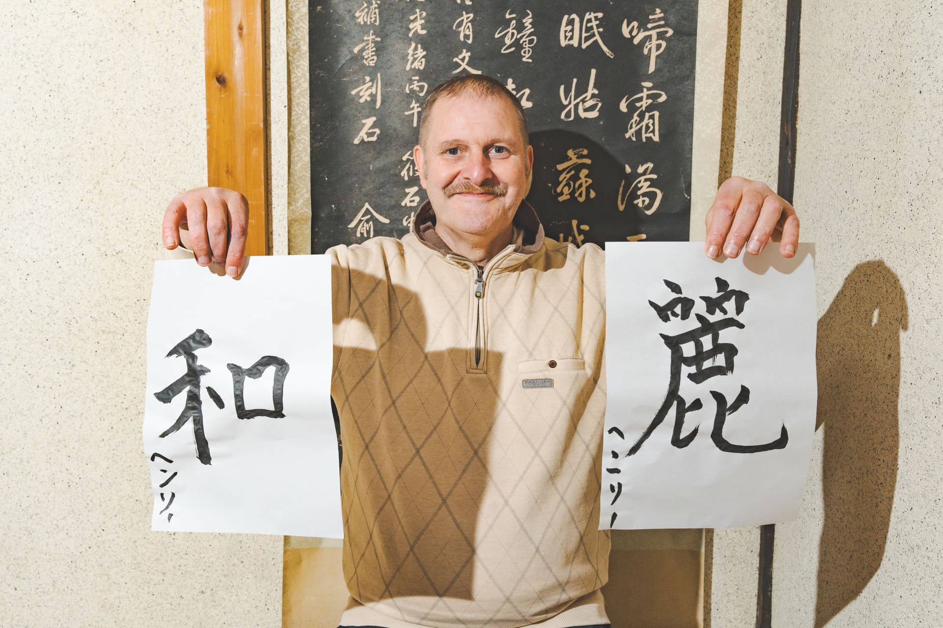 Japanese Cultural Experiences at Kyoto Abeya. A smiling man holding two pieces of calligraphy artwork, with the characters '和' and '愛' written on them, standing in front of a wall with Japanese writing.