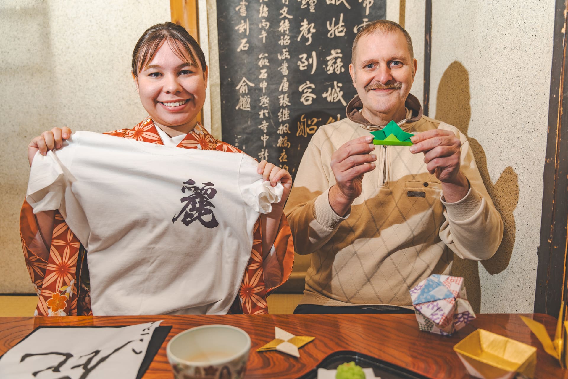 Japanese Cultural Experiences at Kyoto Abeya. A woman in a kimono and a man in a sweater holding a white T-shirt with the character '愛' and a green origami crane, with a blackboard filled with Japanese calligraphy in the background.