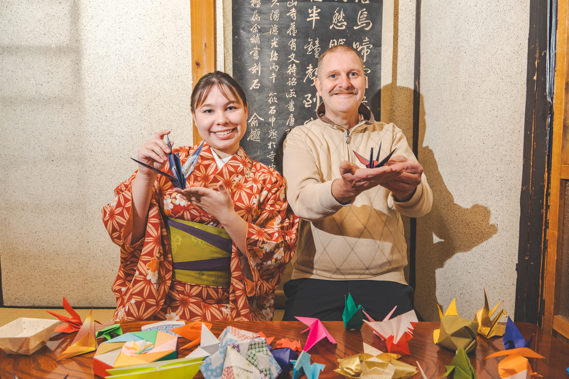 Japanese Cultural Experiences at Kyoto Abeya. A woman in a kimono and a man in a sweater displaying colorful origami birds, with a variety of folded origami figures spread on the table in front of a blackboard with Japanese calligraphy.