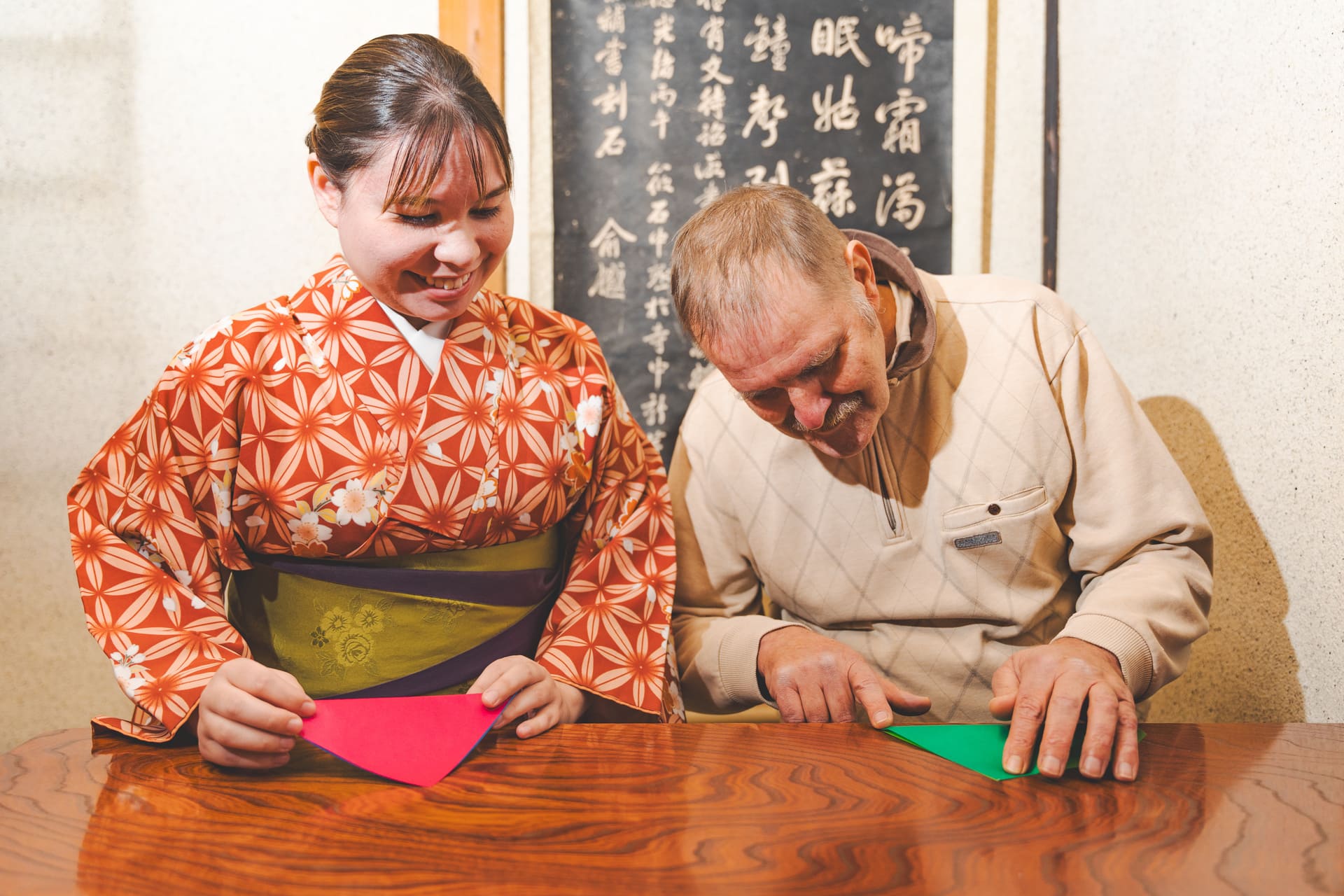 Japanese Cultural Experiences at Kyoto Abeya. A woman in a kimono and a man in a sweater creating origami, focused on their hands as they fold colorful paper, with Japanese calligraphy visible in the background.