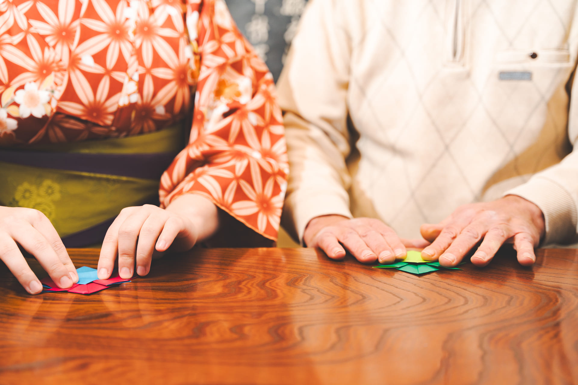 Japanese Cultural Experiences at Kyoto Abeya. The hands of a woman in a kimono and a man in a sweater folding a red and a green piece of origami paper on a wooden table, with a focus on the process of origami folding.