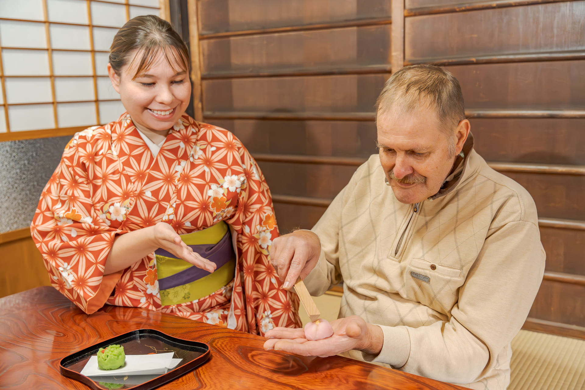 Japanese Cultural Experiences at Kyoto Abeya. A woman in a kimono and a man in a sweater are at a wooden table, with the woman teaching the man how to wrap a Japanese wagashi sweet with a bamboo leaf.