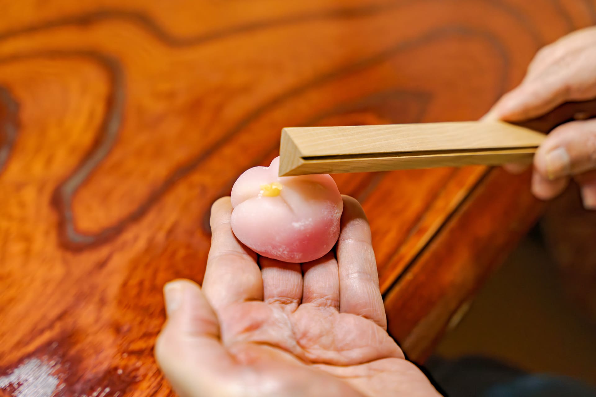 Japanese Cultural Experiences at Kyoto Abeya. Close-up of a man's hands using wooden tongs to pick up a pink wagashi (Japanese sweet), with a wooden table surface.